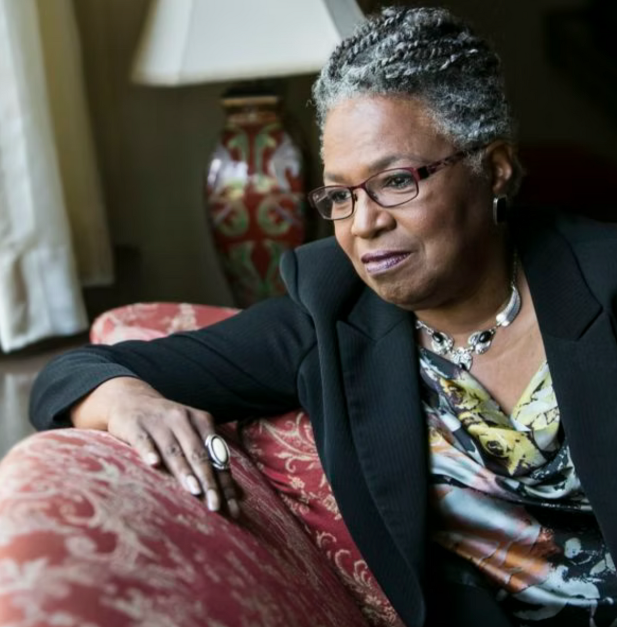 A woman with a crown of gray hair and glasses sits on a red patterned armchair, wearing a black blazer over a multicolor patterned blouse. A decorative lamp is visible in the background.
